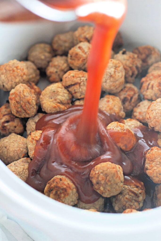 A stream of red sauce being poured over a pile of cooked meatballs in a white dish or slow cooker.
