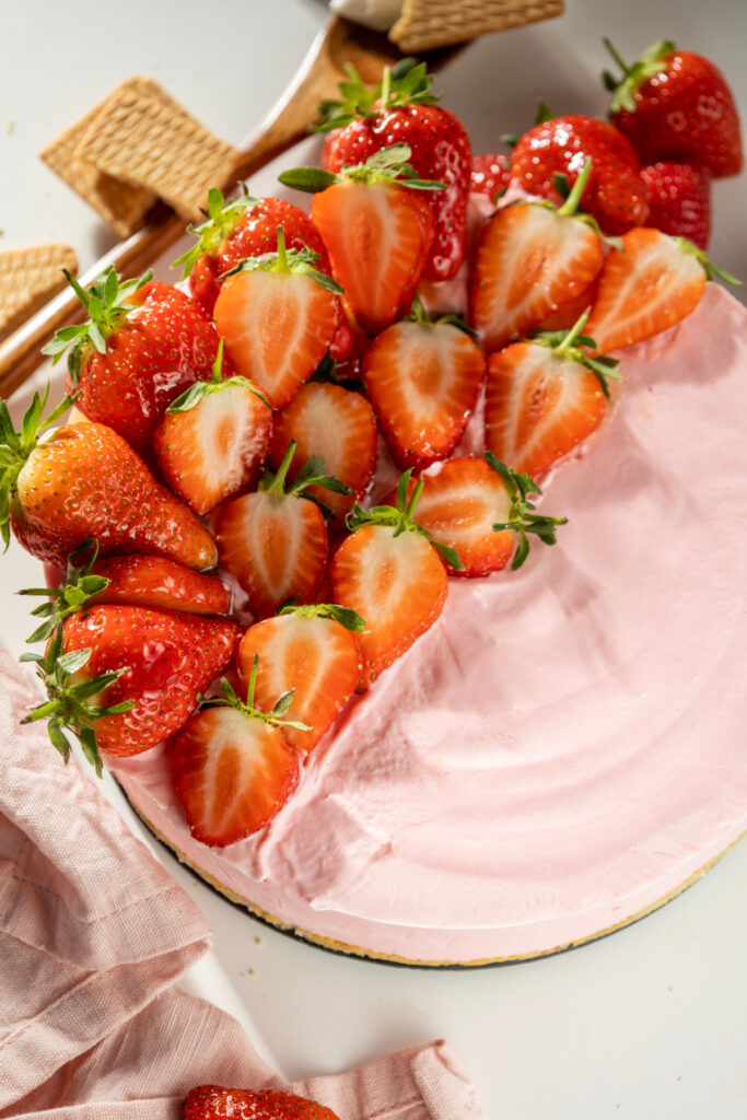 A pink strawberry cheesecake is topped with halved fresh strawberries, with wafer cookies and a pink cloth nearby.