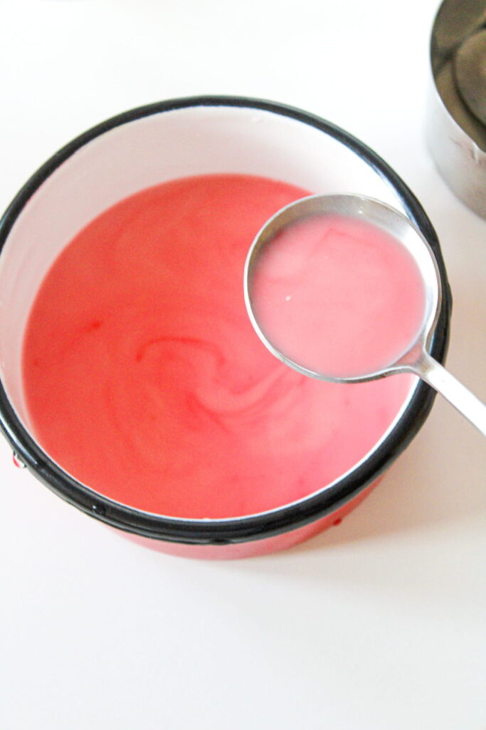 A metal ladle rests above a white bowl with a black rim, filled with a pink liquid.