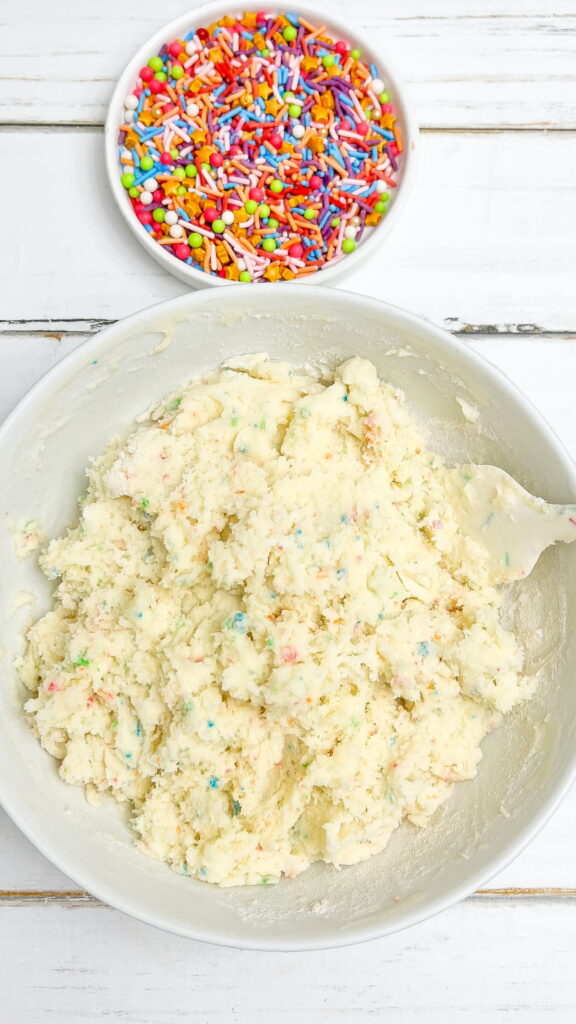 A bowl of funfetti cookie dough with a spatula sits next to a bowl of colorful sprinkles on a white wooden surface.