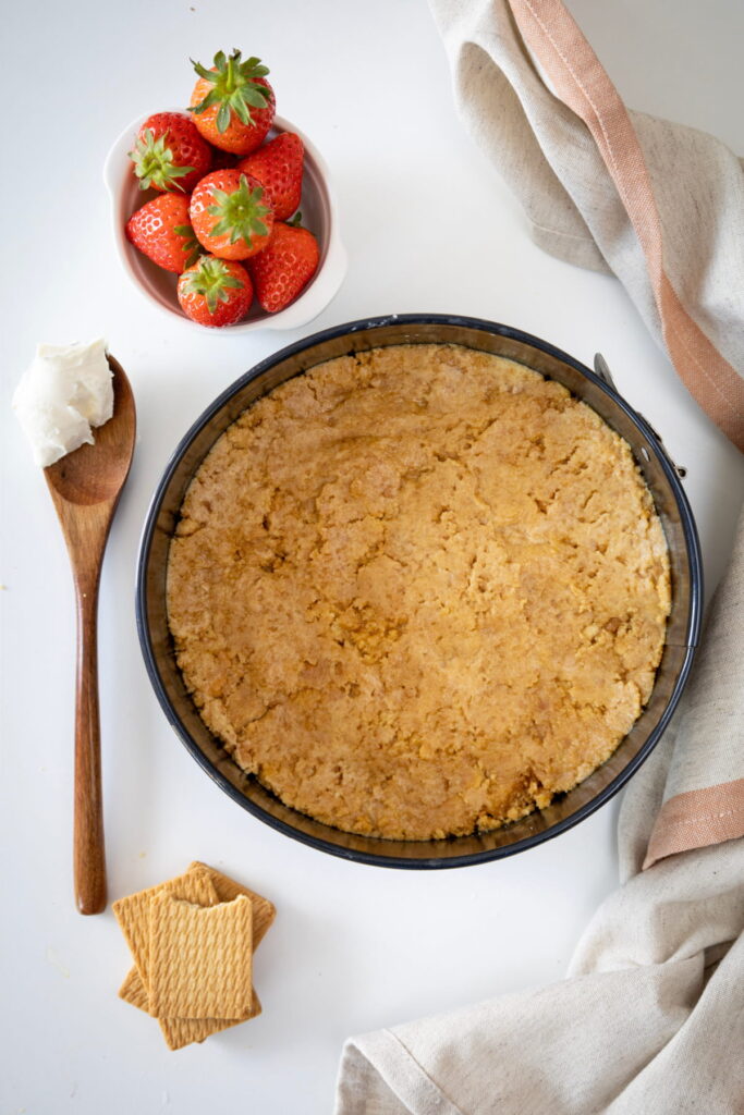 A round baking pan with a prepared crumb crust, a bowl of strawberries, a wooden spoon with a dollop of cream, and square biscuits on a white surface.