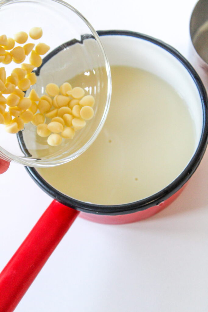 A hand pours white chocolate chips from a glass bowl into a small red saucepan filled with milk.