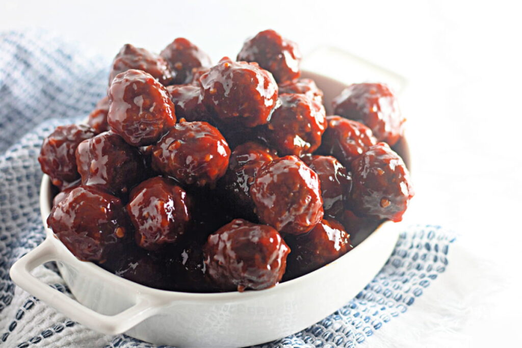 A white dish filled with glazed cocktail meatballs, set on a textured blue and white cloth.