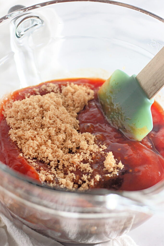 A glass mixing bowl with ketchup, brown sugar, and a green spatula, ready for mixing.