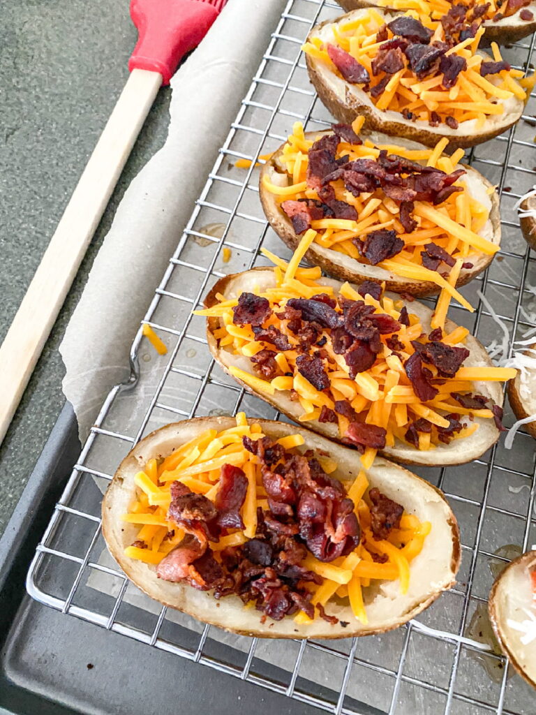 Baked potato skins topped with shredded cheddar cheese and crispy bacon bits on a wire rack over a baking tray, with a red spatula nearby.