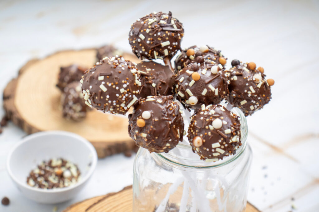 Chocolate cake pops coated in sprinkles are arranged on sticks in a glass jar, with more treats and a bowl of sprinkles visible in the background.