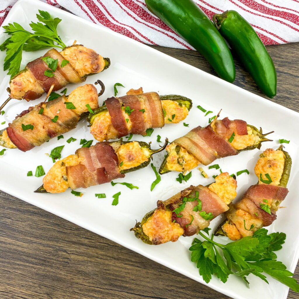 A white plate with six bacon-wrapped stuffed jalapeño halves, garnished with chopped parsley, on a wooden table beside whole jalapeños and a red-striped napkin.