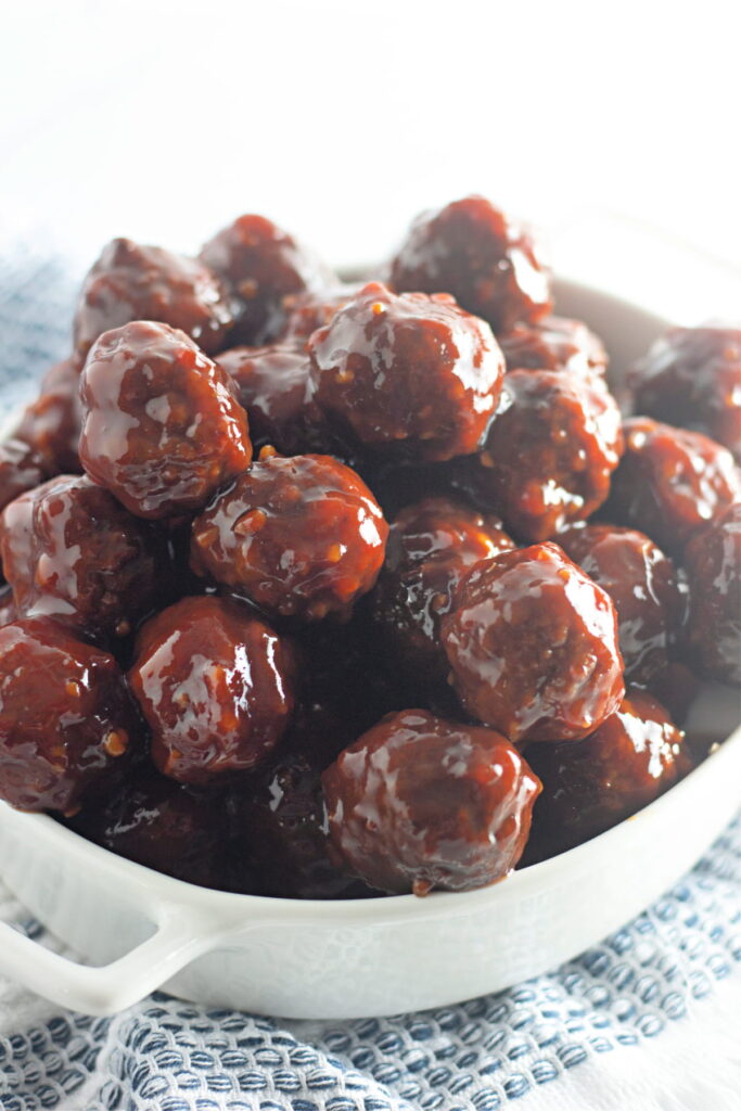 A white dish filled with glazed meatballs, piled high and coated in a shiny, dark sauce, resting on a blue and white patterned cloth.