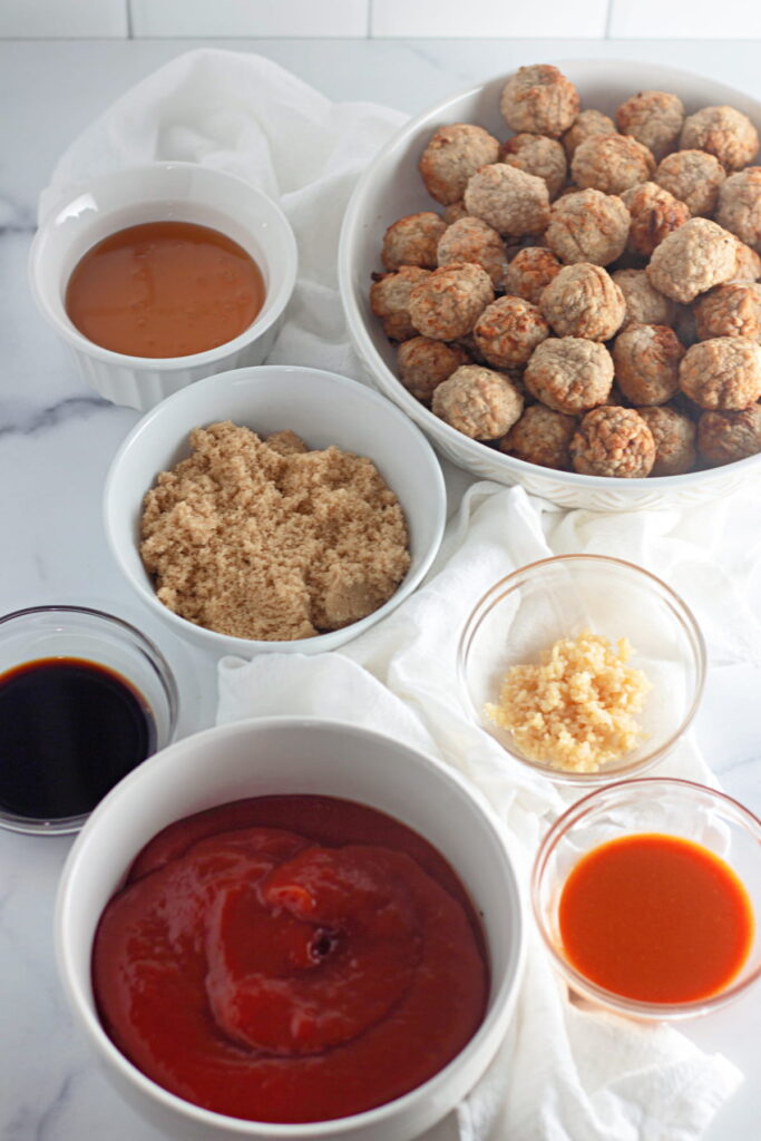A variety of ingredients in bowls on a white surface, including cooked meatballs, brown sugar, tomato sauce, soy sauce, minced garlic, hot sauce, and another sauce.