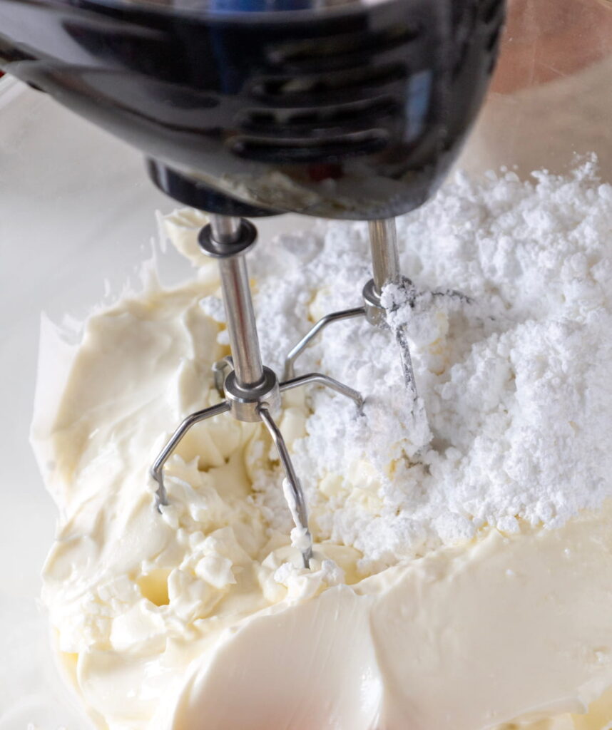 Electric hand mixer beaters in a mixing bowl with cream cheese and powdered sugar.