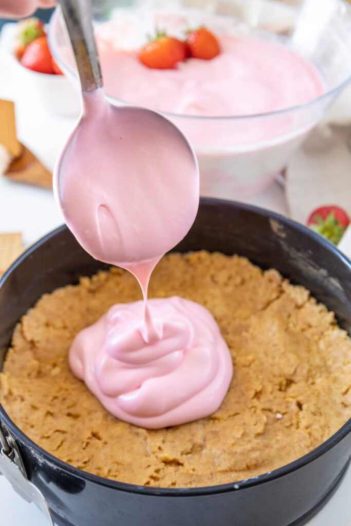 A spoon is pouring pink strawberry mousse onto a graham cracker crust in a springform pan, with more mousse and strawberries in the background.