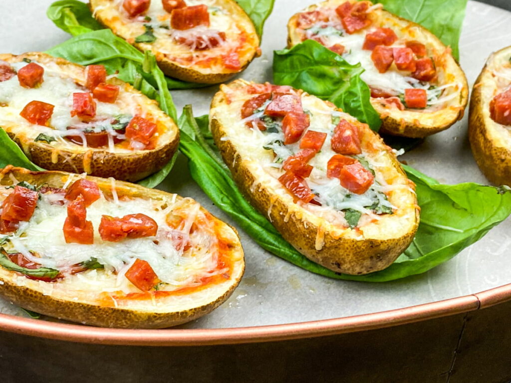 Potato skins topped with melted cheese, diced peppers, and herbs, served on a metal tray with fresh green leaves underneath each piece.
