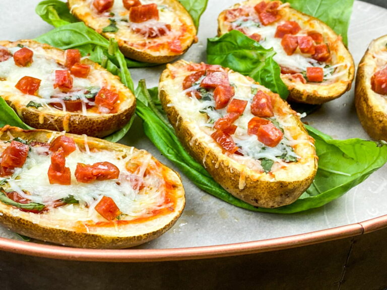 Potato skins topped with melted cheese, diced peppers, and herbs, served on a metal tray with fresh green leaves underneath each piece.