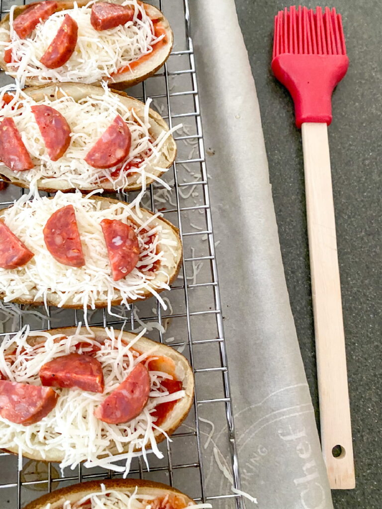 Potato skins topped with shredded cheese and pepperoni on a wire rack lined with parchment paper, next to a red silicone basting brush.