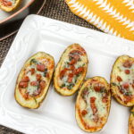 Four potato skins on a white plate, filled with melted cheese, chopped pepperoni, and herbs, with a yellow patterned napkin beside the plate.