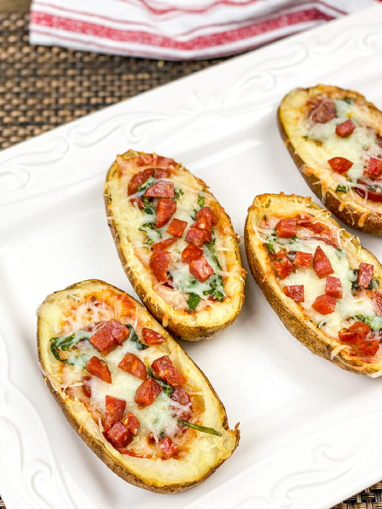 Four potato skins topped with melted cheese, diced tomatoes, and herbs are arranged on a white rectangular plate.