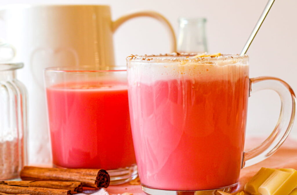 Two clear mugs filled with pink-hued drinks, one topped with foam, sit on a table with cinnamon sticks and glass containers nearby.