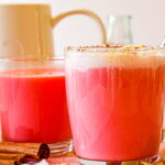 Two clear mugs filled with pink-hued drinks, one topped with foam, sit on a table with cinnamon sticks and glass containers nearby.