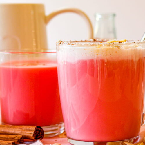 Two clear mugs filled with pink-hued drinks, one topped with foam, sit on a table with cinnamon sticks and glass containers nearby.