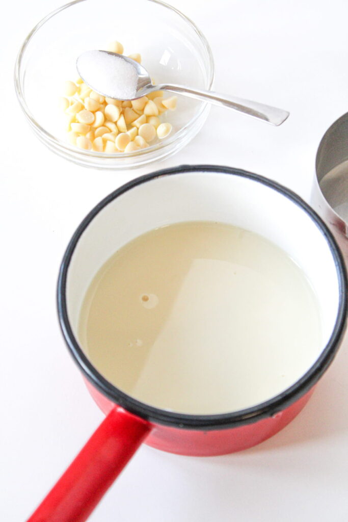 A red saucepan with liquid, a bowl of white chocolate chips, and a spoon with white powder, likely sugar, on a white surface.