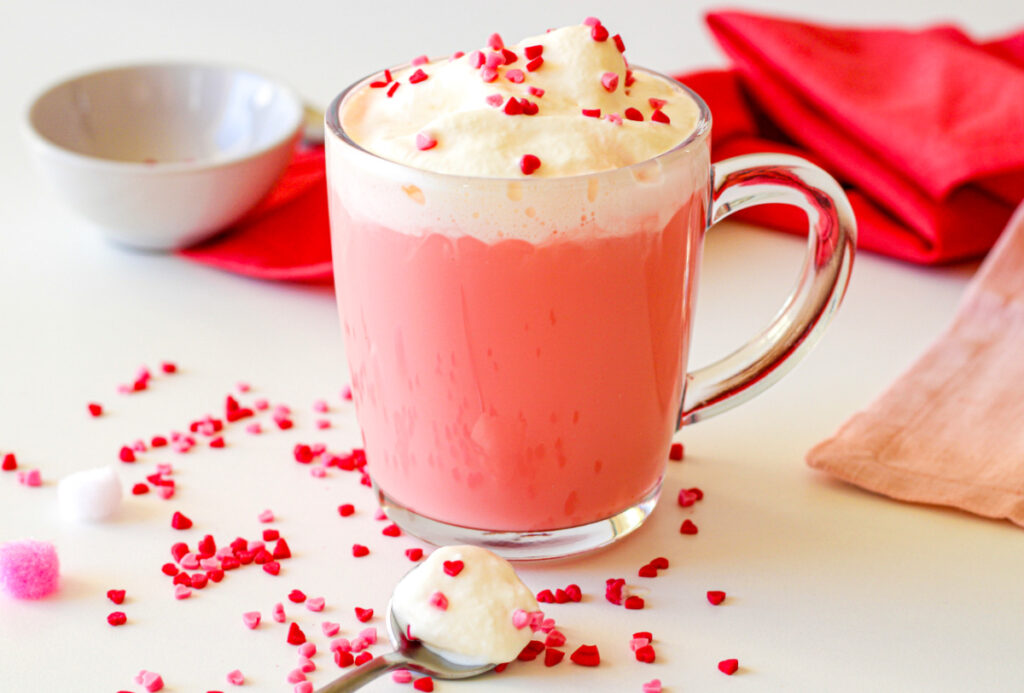A glass mug of pink beverage topped with whipped cream and red heart sprinkles, with more sprinkles on the table and a spoon in the foreground.