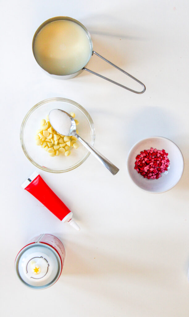 A measuring cup of milk, a bowl of white chocolate chips with a spoonful of sugar, a bowl of pink heart sprinkles, red gel tube, and a can of whipped cream on a white surface.