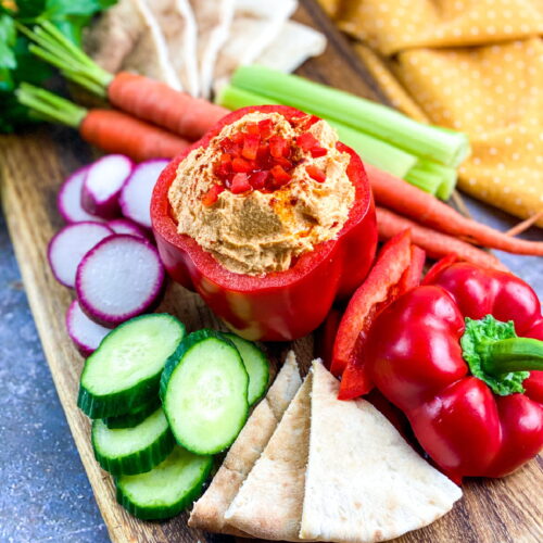 A wooden board with pita bread, sliced cucumber, radish, carrot, celery, and a red bell pepper filled with hummus, garnished with diced red pepper.