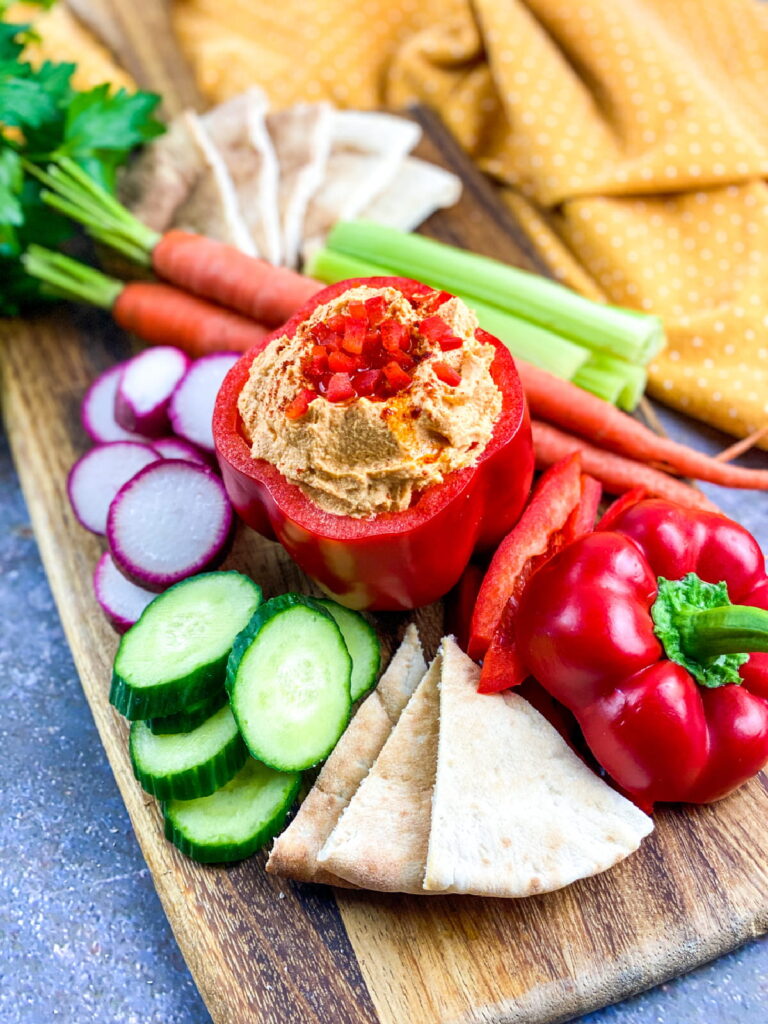 A wooden board with pita bread, sliced cucumber, radish, carrot, celery, and a red bell pepper filled with hummus, garnished with diced red pepper.