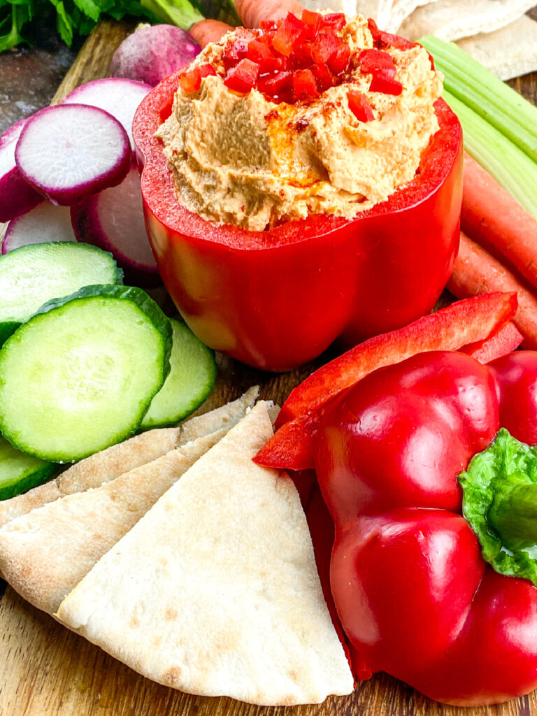 Red bell pepper filled with hummus, topped with diced red peppers, surrounded by sliced cucumber, radish, carrots, pita wedges, and another whole red bell pepper on a wooden board.