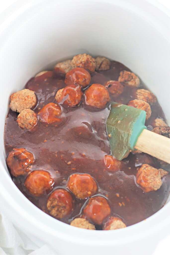 Meatballs in a thick brown sauce inside a white slow cooker, with a green spatula partially submerged in the mixture.
