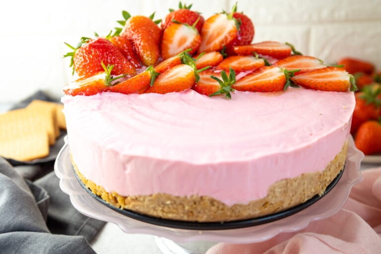 A pink strawberry cheesecake with a biscuit base, topped with halved fresh strawberries, displayed on a glass cake stand.
