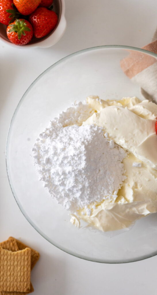 A glass bowl containing cream cheese and powdered sugar, with graham crackers and strawberries nearby on a white surface.