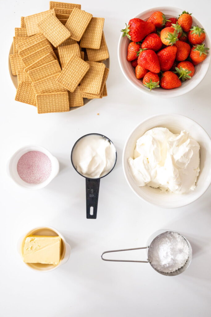 Top-down view of cheesecake ingredients: rectangular biscuits, fresh strawberries, cream cheese, sour cream, butter, powdered sugar, and pink powder, each in separate bowls or containers.