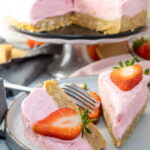 A sliced pink strawberry cheesecake with a cookie crust, garnished with fresh strawberries, served on a plate with a fork.