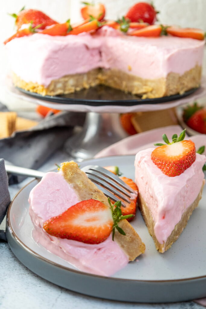 A sliced pink strawberry cheesecake with a cookie crust, garnished with fresh strawberries, served on a plate with a fork.