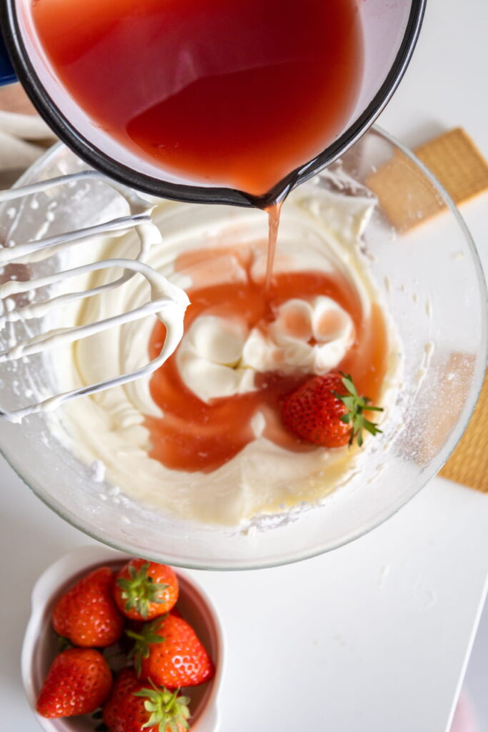 Strawberry syrup is being poured into a bowl of whipped cream with strawberries, next to a small bowl of whole strawberries on a white surface.
