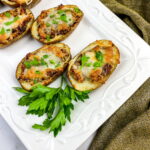 Three twice-baked potato halves topped with melted cheese and herbs are arranged on a white plate, garnished with fresh parsley. A green cloth lies beside the plate.