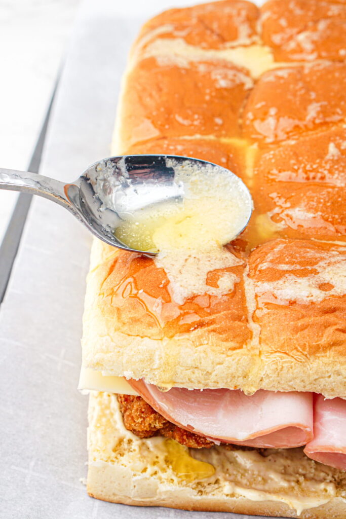 A spoon drizzles melted butter over a sandwich made with layered ham, cheese, and breaded chicken on a tray of sliced dinner rolls.