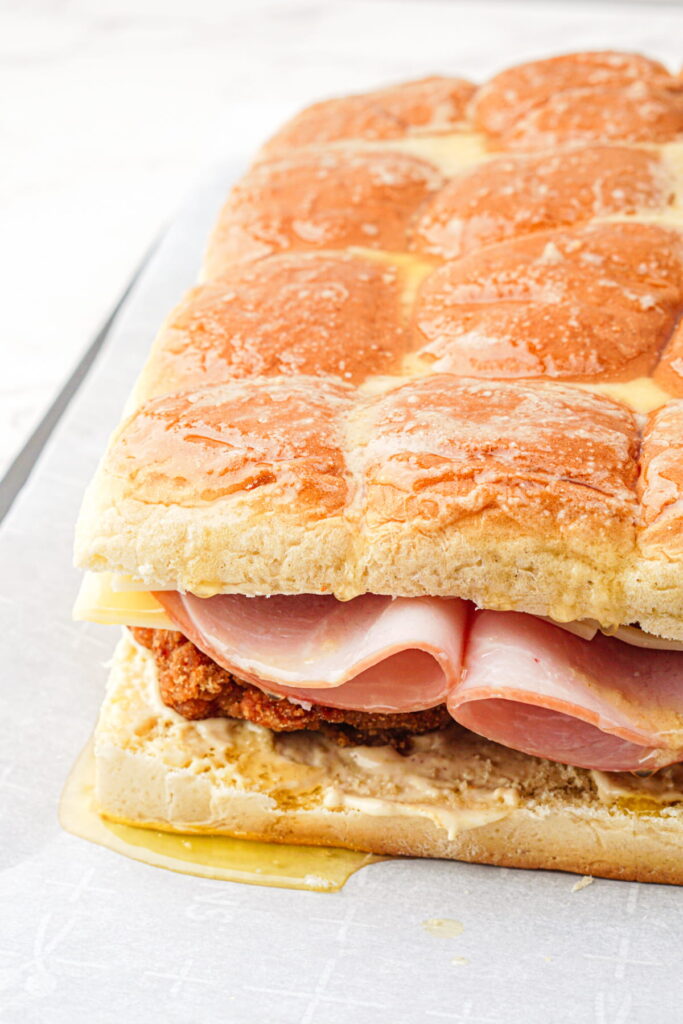 A close-up of a large sandwich made with rolls, layered with ham, cheese, and breaded chicken, with melted butter visible on the bread.