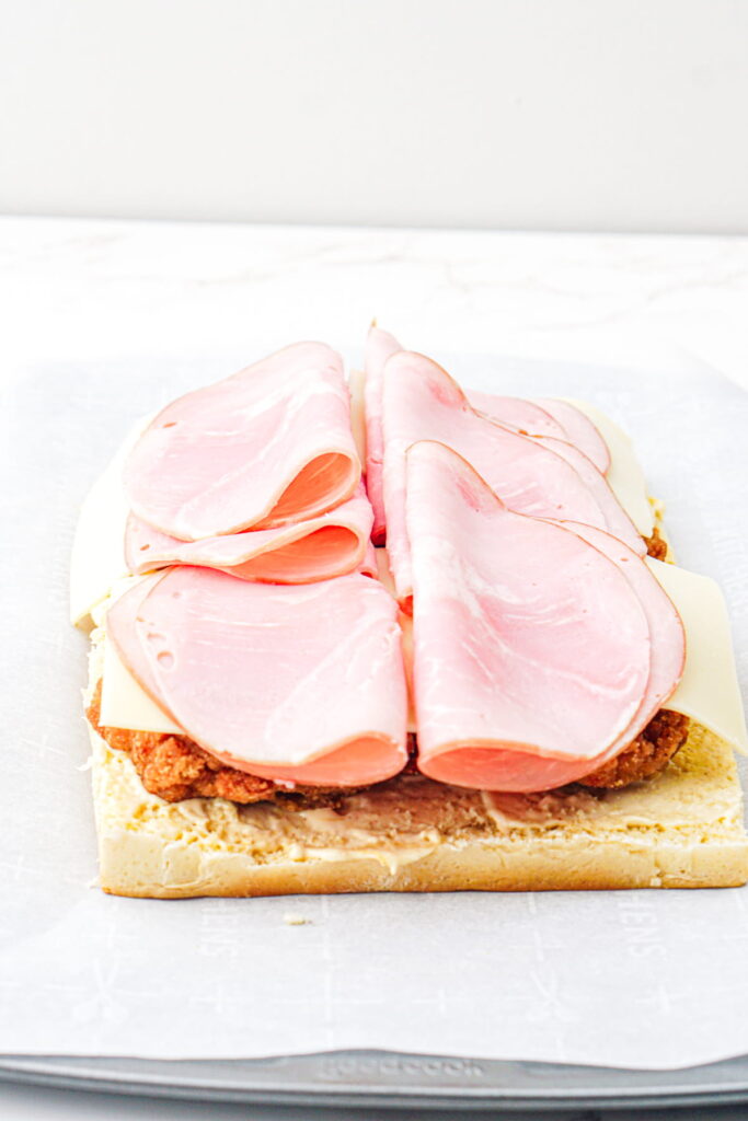 An open sandwich with layers of ham, cheese slices, and a breaded meat patty on a rectangular piece of bread.