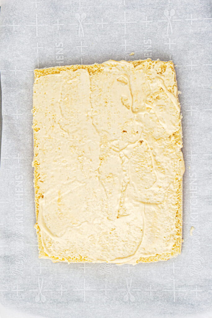 A rectangular sponge cake layer topped with an even spread of cream on parchment paper.