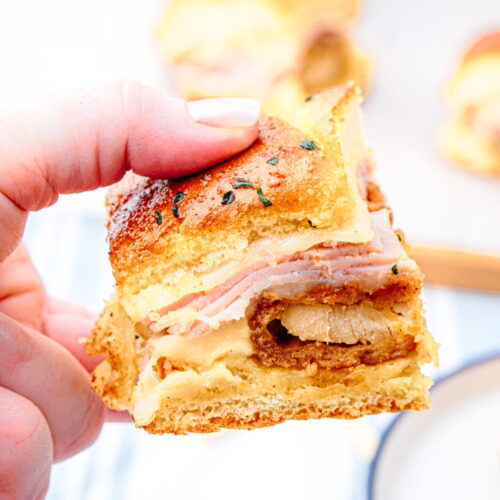 A hand holding a small sandwich with layers of sliced ham, cheese, and a piece of breaded meat between golden bread slices.