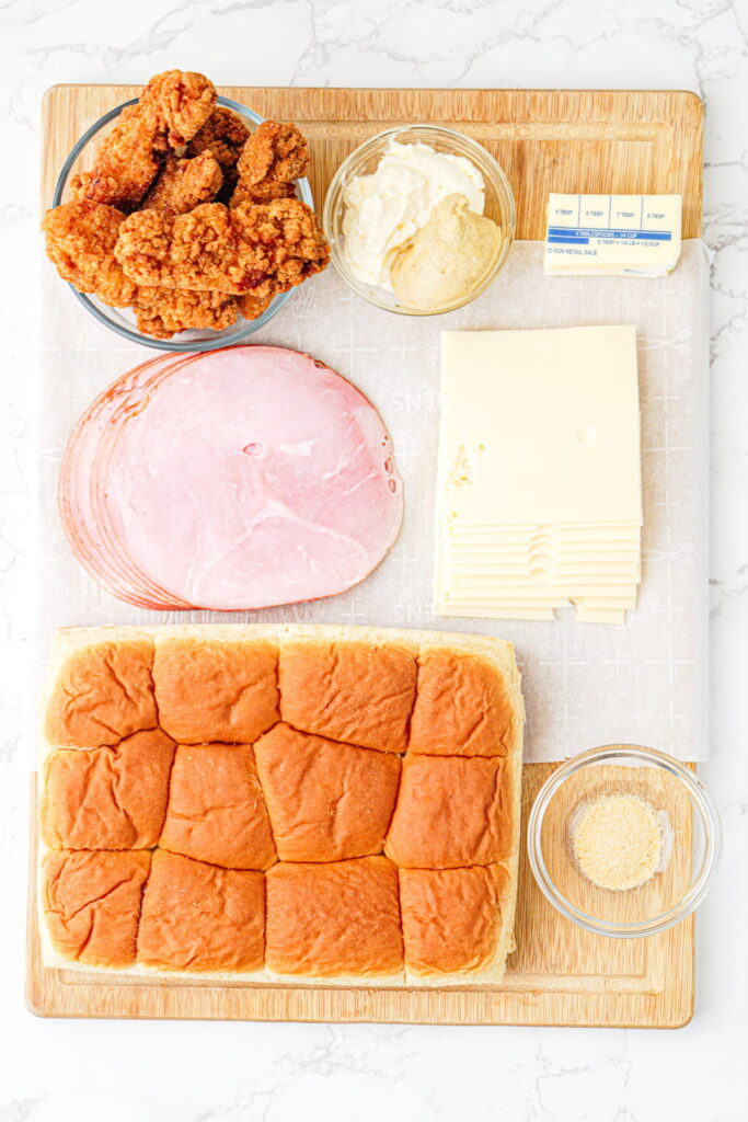 Ingredients on a cutting board, including fried chicken, ham, Swiss cheese slices, buns, mayonnaise, mustard, butter, and a bowl of breadcrumbs.