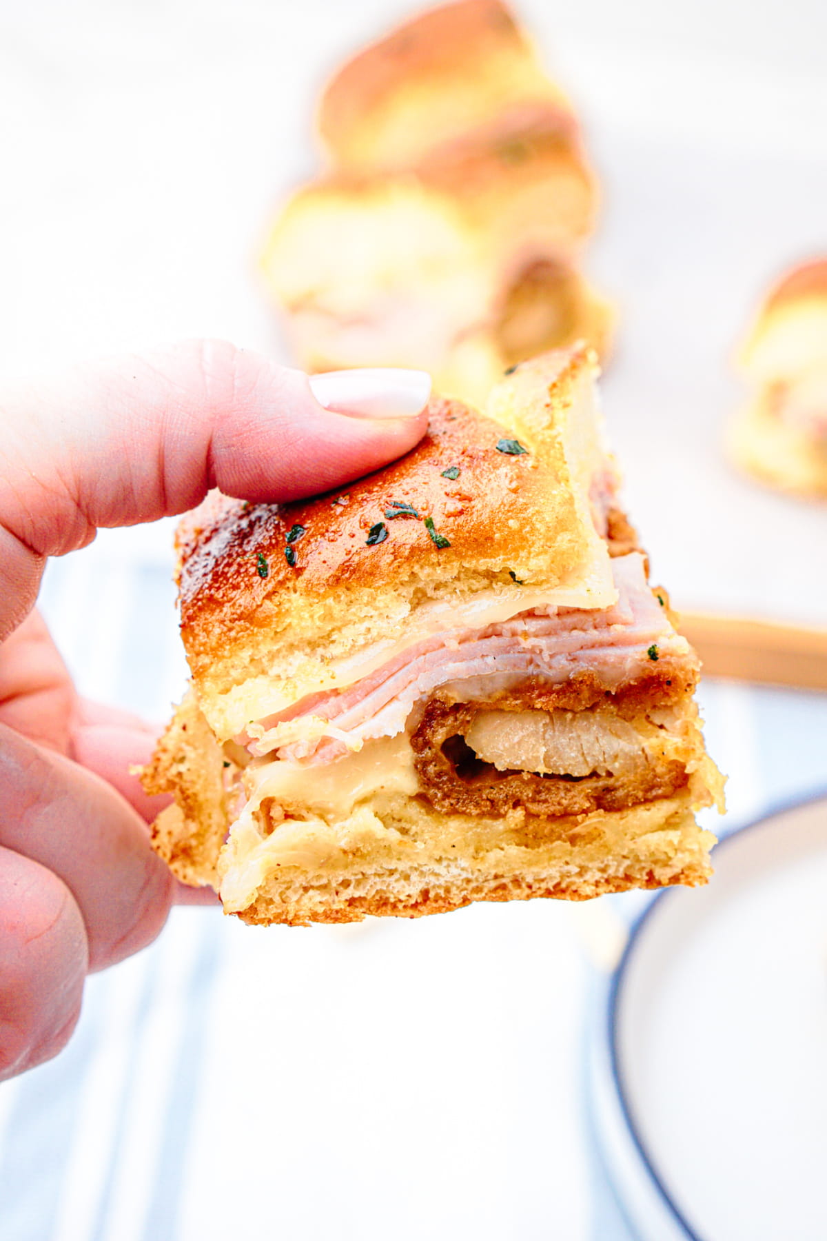 A hand holding a small sandwich with layers of sliced ham, cheese, and a piece of breaded meat between golden bread slices.