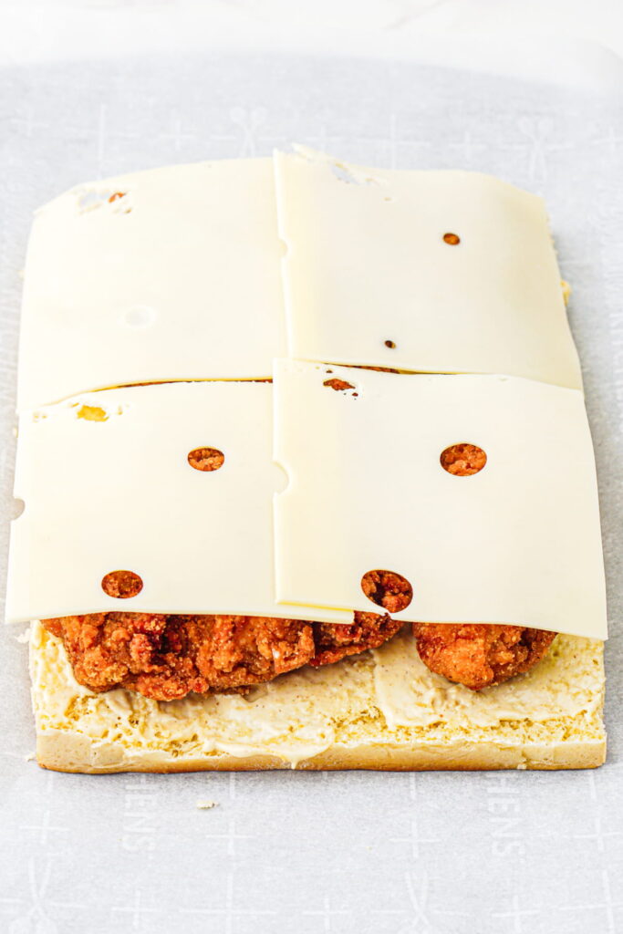 Four slices of Swiss cheese are placed on top of breaded chicken pieces on the bottom half of a sandwich roll.