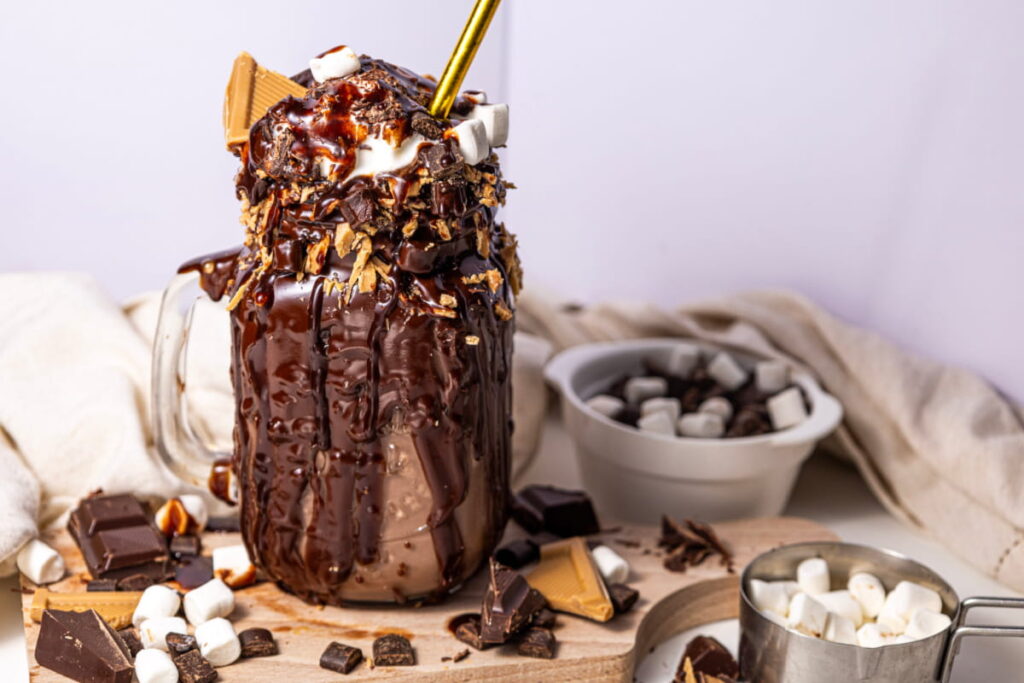 A mason jar filled with chocolate milkshake, topped with chocolate sauce, marshmallows, and graham crackers, sits on a board with scattered chocolate pieces and marshmallows nearby.