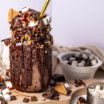 A mason jar filled with chocolate milkshake, topped with chocolate sauce, marshmallows, and graham crackers, sits on a board with scattered chocolate pieces and marshmallows nearby.