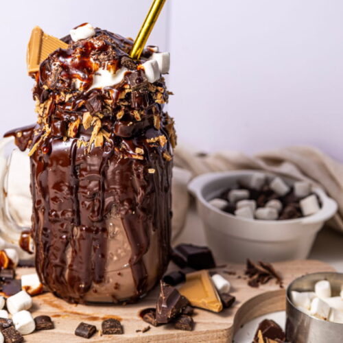 A mason jar filled with chocolate milkshake, topped with chocolate sauce, marshmallows, and graham crackers, sits on a board with scattered chocolate pieces and marshmallows nearby.