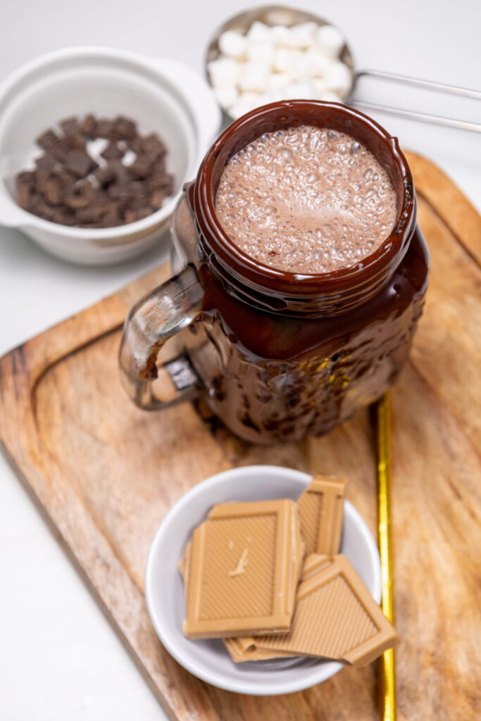 A mason jar mug filled with frothy chocolate drink sits on a wooden board, surrounded by white chocolate squares, chocolate chunks, and a bowl of sugar cubes.