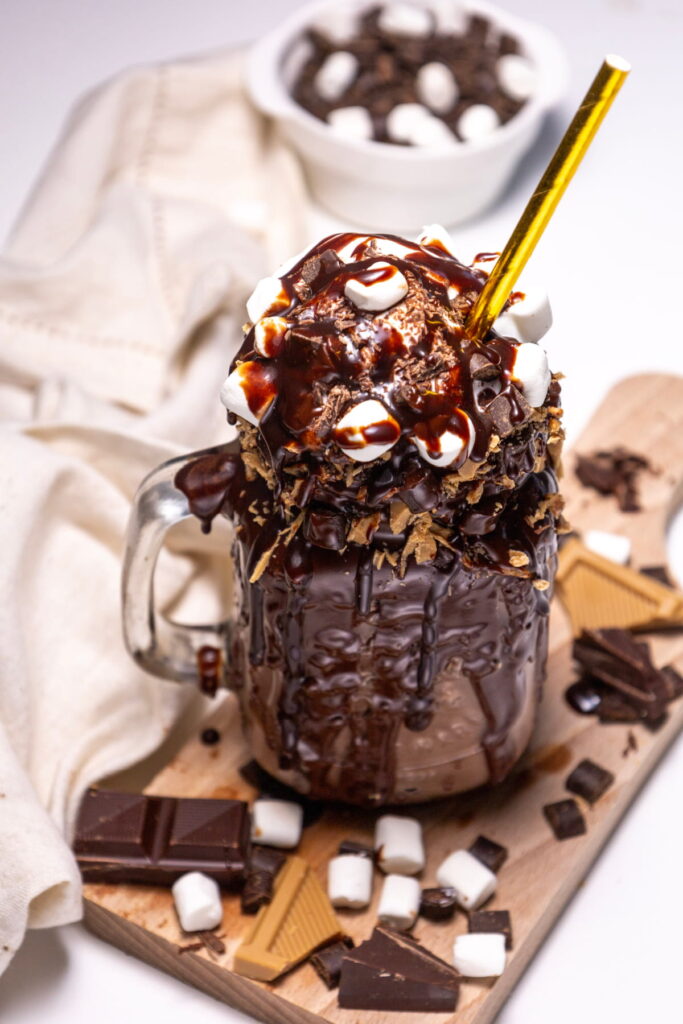 A glass mug filled with a chocolate milkshake, topped with whipped cream, chocolate syrup, marshmallows, and a gold straw, surrounded by chocolate pieces on a wooden board.